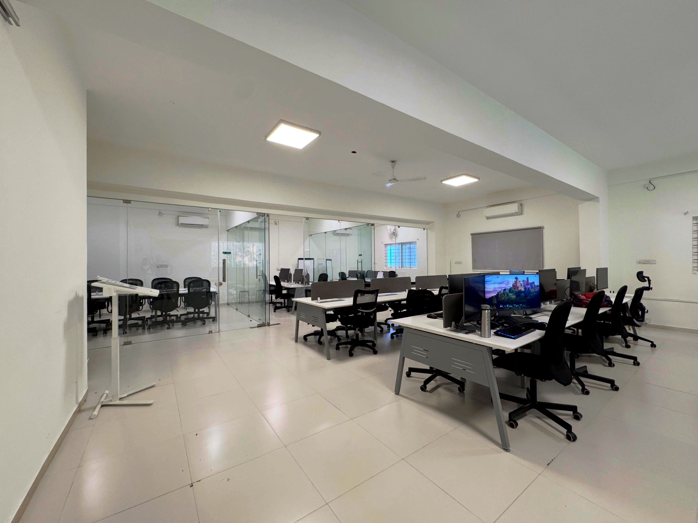 Open-plan floor of the JoinNow Bangalore office with rows of white desks, mesh task chairs and glass-partitioned team rooms.